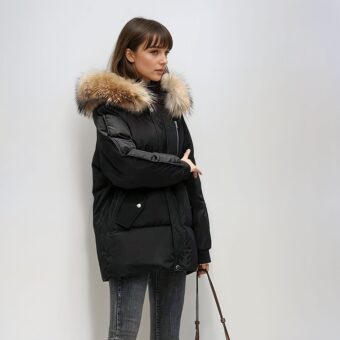 Bruinharige vrouw met franje, staand voor een wit/grijze muur, draagt een zwart donsjack met capuchon en bruin gewatteerd bont, met een handtasriem in haar linkerhand.