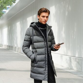 Halflange donsjas voor mannen in grijs, gedragen door een jonge man met bruin haar, die zijn telefoon in zijn linkerhand houdt. Hij staat op straat.