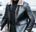 We zien het borstbeeld van een man die over straat loopt, een zwarte trui en een leren jas met een bontkraag draagt. Hij ziet er elegant uit.