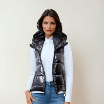 Een brunette vrouw met een blauwe spijkerbroek, een witte top met lange mouwen en een mouwloos gewatteerd donsjack met een glanzende donkergrijze capuchon, die met haar rug naar een beige muur staat.