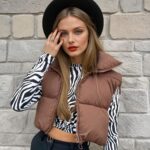 Een vrouw met lang blond haar draagt een zwarte broek, een kort topje met lange mouwen met een wit en zwart zebrapatroon, een bruin gewatteerd mouwloos jack en een zwarte hoed. Ze staat met haar rug naar een beige stenen muur.