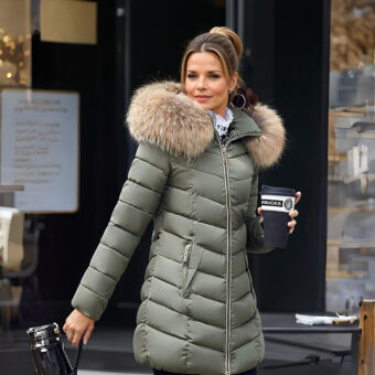 Vrouw staat op een stoep en draagt een kaki groen gewatteerd donsjack met bontmuts, houdt een zwarte beker in haar linkerhand en een zwarte handtas in haar rechterhand.