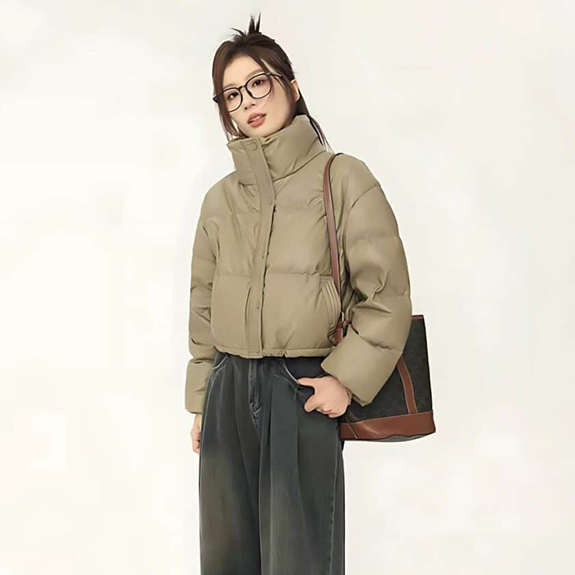 Aziatische vrouw met achterover gebonden haar en bril. Ze draagt een zwarte broek en een kort kakikleurig kunstlederen jasje. Haar handen zitten in de zakken van haar broek. Op haar linkerschouder draagt ze een zwart-bruine handtas. Op een beige achtergrond.