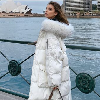 Een bruinharige vrouw staat links van een groene metalen barrière. Rechts van de barrière is een stuk rivierachtig water met gebouwen op de achtergrond. De vrouw draagt een lange witte donsjas met een witte bontmuts. In haar linkerhand heeft ze een handtasriem. Ze is in profiel.