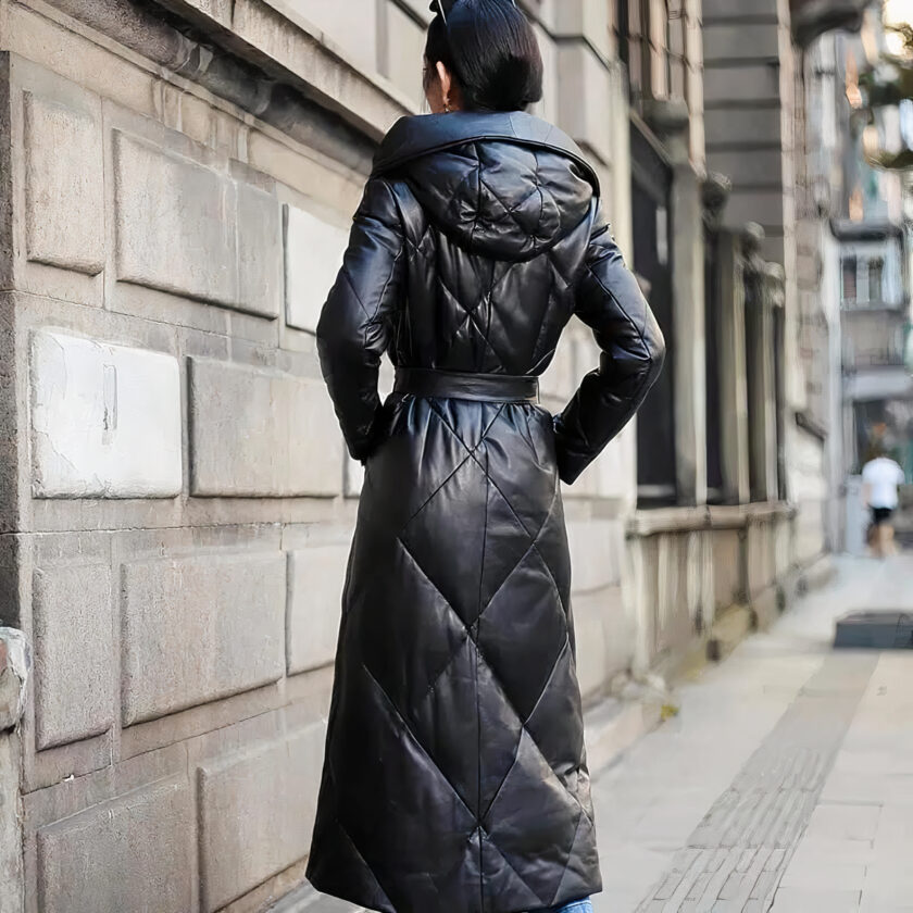 We zien een donkerharige vrouw van achteren, op straat. Ze draagt een knotje. Ze draagt een lange zwarte imitatieleren jas. Ze heeft haar handen in haar zakken. Ze staat naast een hele mooie muur, typisch voor Parijs, of van een grote stad met mooie architectuur.