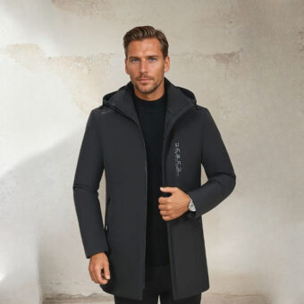 Een man met kort, lichtbruin haar, ongeschoren, maar met een getailleerde zwarte jas met capuchon. Zijn linkerhand houdt de rits rond zijn middel. Op een beige achtergrond.