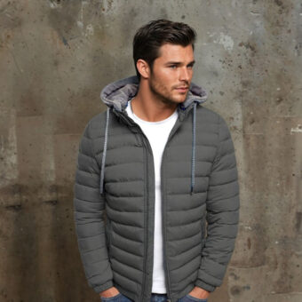 Een donkerharige man met een ongeschoren baard staat voor een donkergrijze muur, draagt een wit T-shirt, een blauwe spijkerbroek en een kort, dun, gewatteerd grijs donsjack met capuchon en trekkoord. Zijn handen zitten in zijn broekzakken.