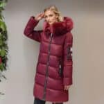 Blonde vrouw in profiel naast een lichtbruine muur met een warme bordeauxrode halflange donsjas met bijpassend bont op de capuchon en zijzakken. Ze houdt haar rechterhand tegen haar slaap. Links is een groene plant te zien.