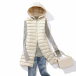 Fijne lange beige mouwloze donsjas met capuchon, plat gepresenteerd op een witte achtergrond met alle accessoires voor een complete outfit.