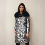 Bruine vrouw met haar rug naar een beige muur, draagt een zwarte broek en een glanzend grijs halflang donsjack met zwart bont op de capuchon. Twee vierkante zijzakken.