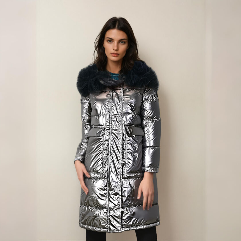 Bruine vrouw met haar rug naar een beige muur, draagt een zwarte broek en een glanzend grijs halflang donsjack met zwart bont op de capuchon. Twee vierkante zijzakken.