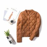 Het donsjack in bomberstijl voor dames is afgebeeld op een witte vloer met een café, documenten en een cactus.