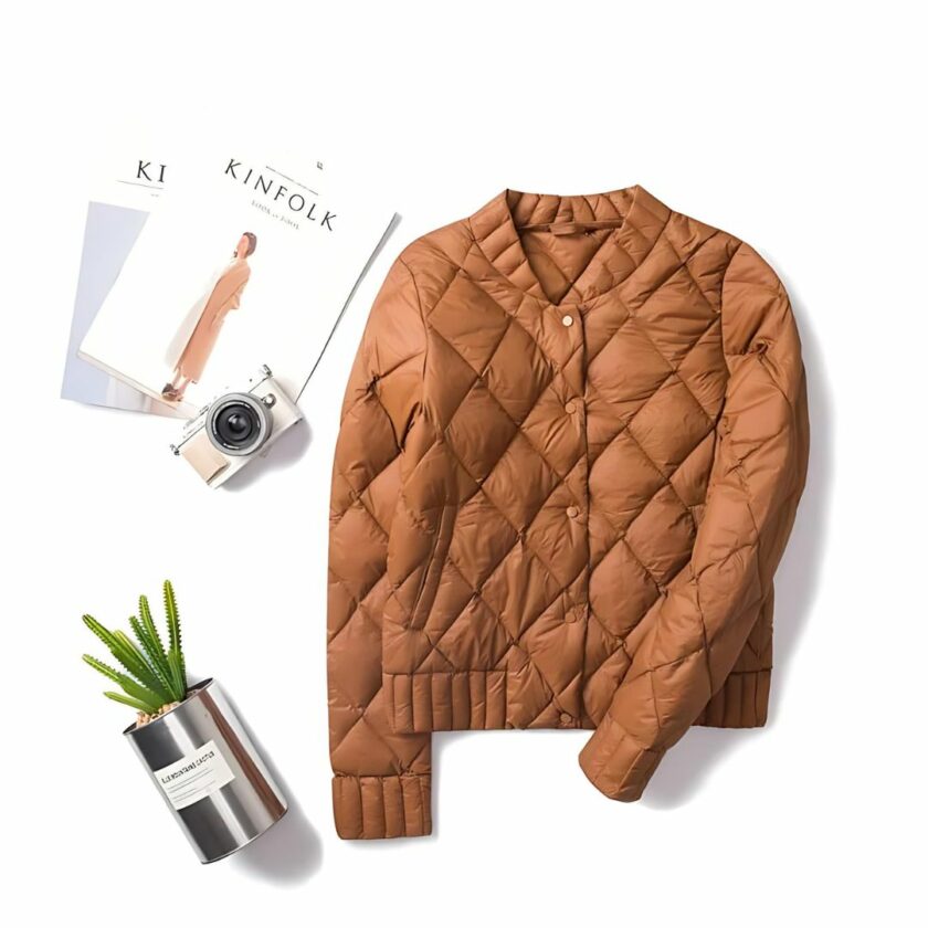 Het donsjack in bomberstijl voor dames is afgebeeld op een witte vloer met een café, documenten en een cactus.