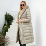 Vrouw draagt een lange gewatteerde beige donsjas met beige bontkraag, vastgemaakt met een riem. Ze heeft lang los haar en een zonnebril met een zwarte tas in haar hand met een witte trap op de achtergrond.