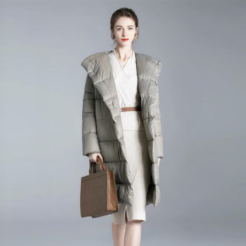 Tegen een grijze achtergrond draagt een vrouw met achterover gebonden bruin haar en mooie oorbellen een lange, loszittende grijze donsjas met een bruine tas in haar rechterhand. Ze draagt een knielange beige jurk.